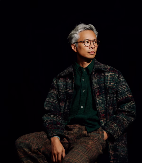 Man with gray hair and glasses wearing a dark green shirt and plaid jacket, seated against a black background.